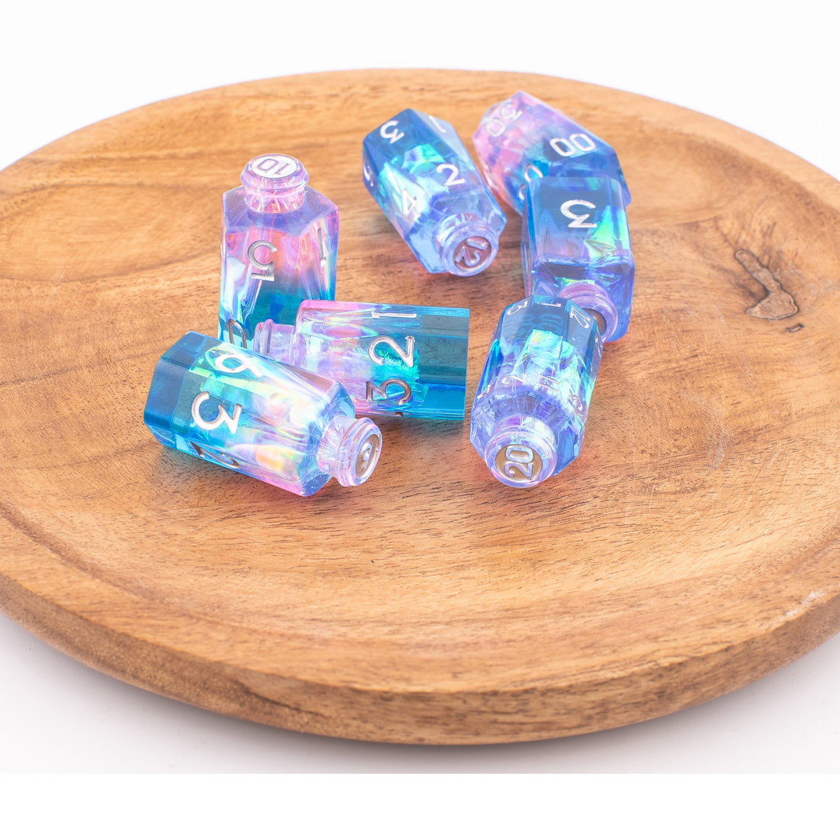 Tabletop Dominion Prismatic Elixir potion bottle dice displayed loose on a wooden tray with iridescent blue‑pink‑purple resin and white numbering.