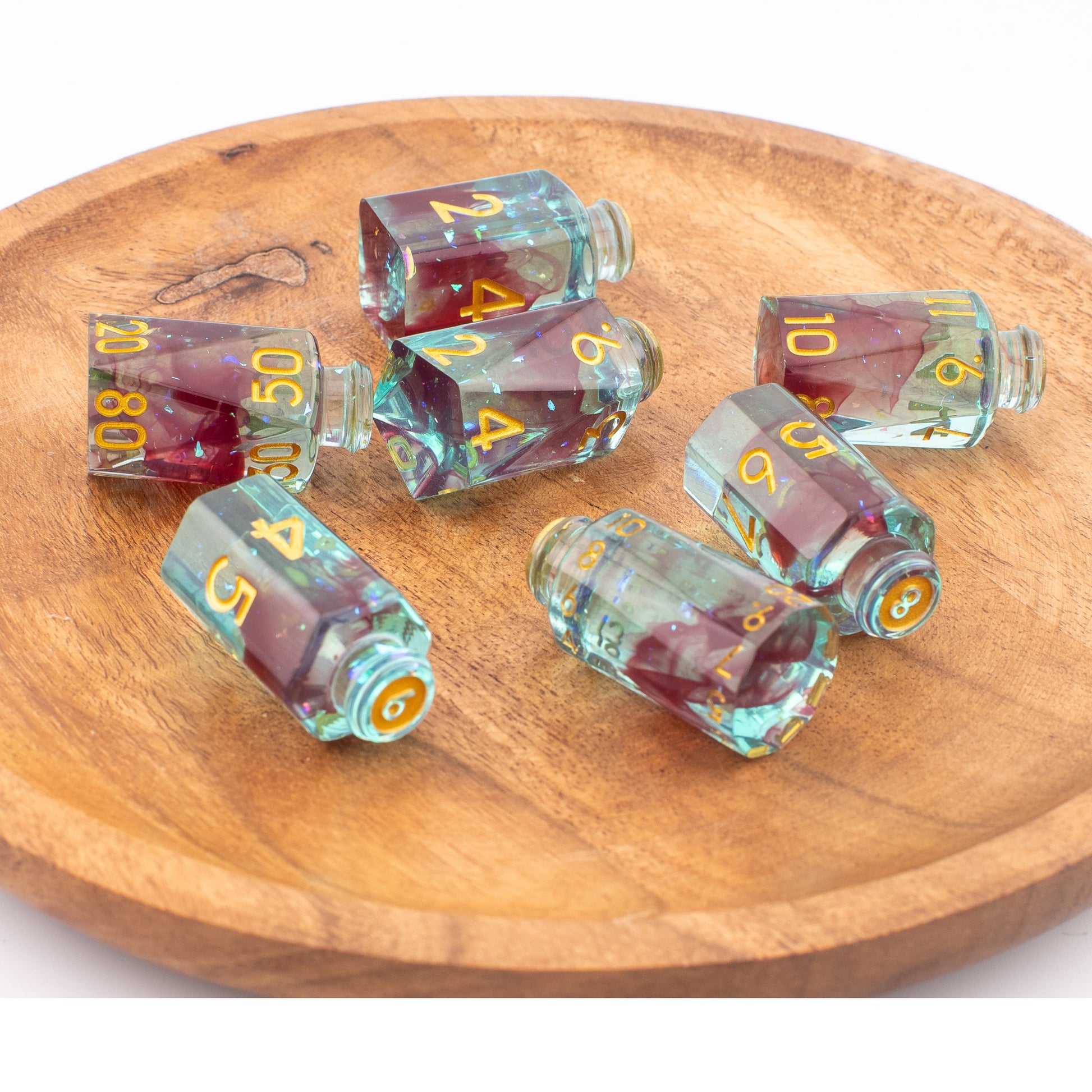 Tabletop Dominion Blood Alchemist potion bottle dice displayed loose on a wooden tray, showing translucent turquoise dice with red inclusions and gold numbering.