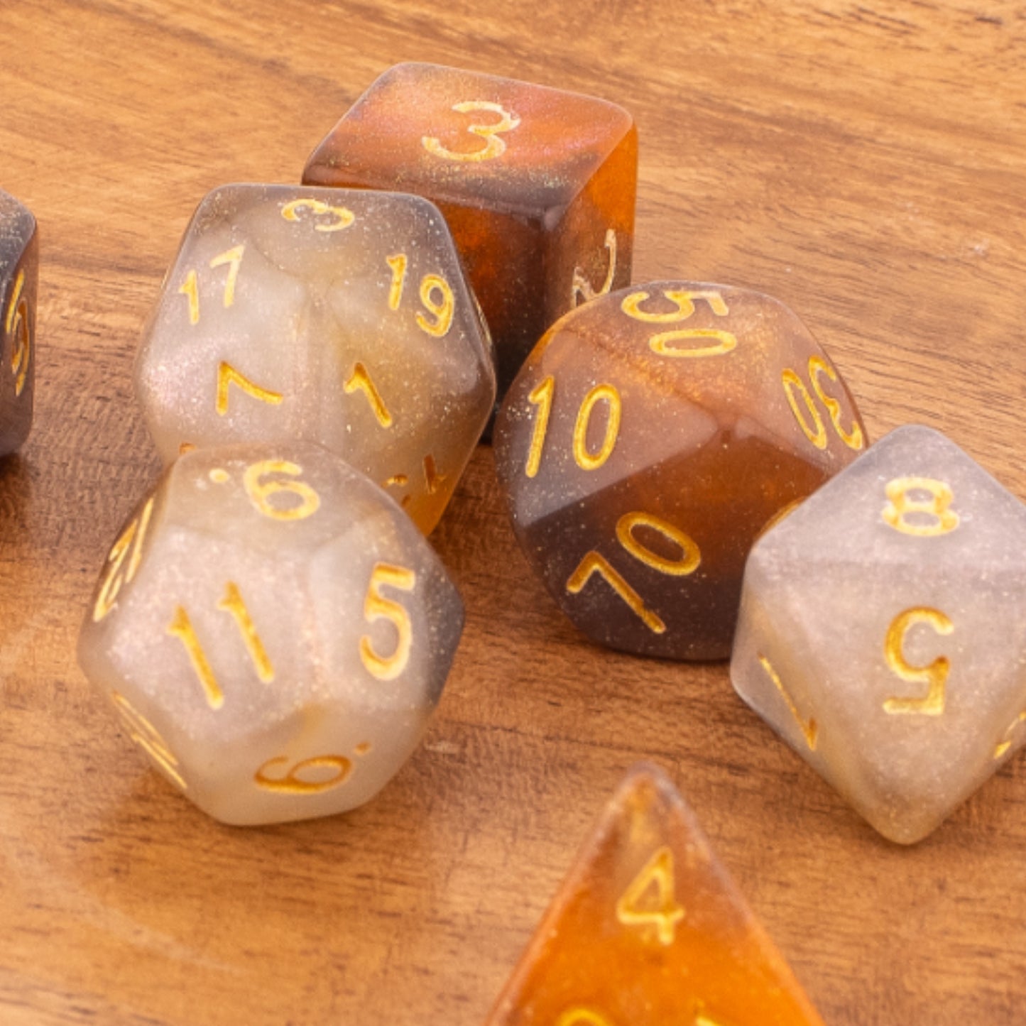 Clear Autumn acrylic dice scattered on a wooden surface, contrasting the warm wood grain with the translucent orange‑grey dice and gold numbering.