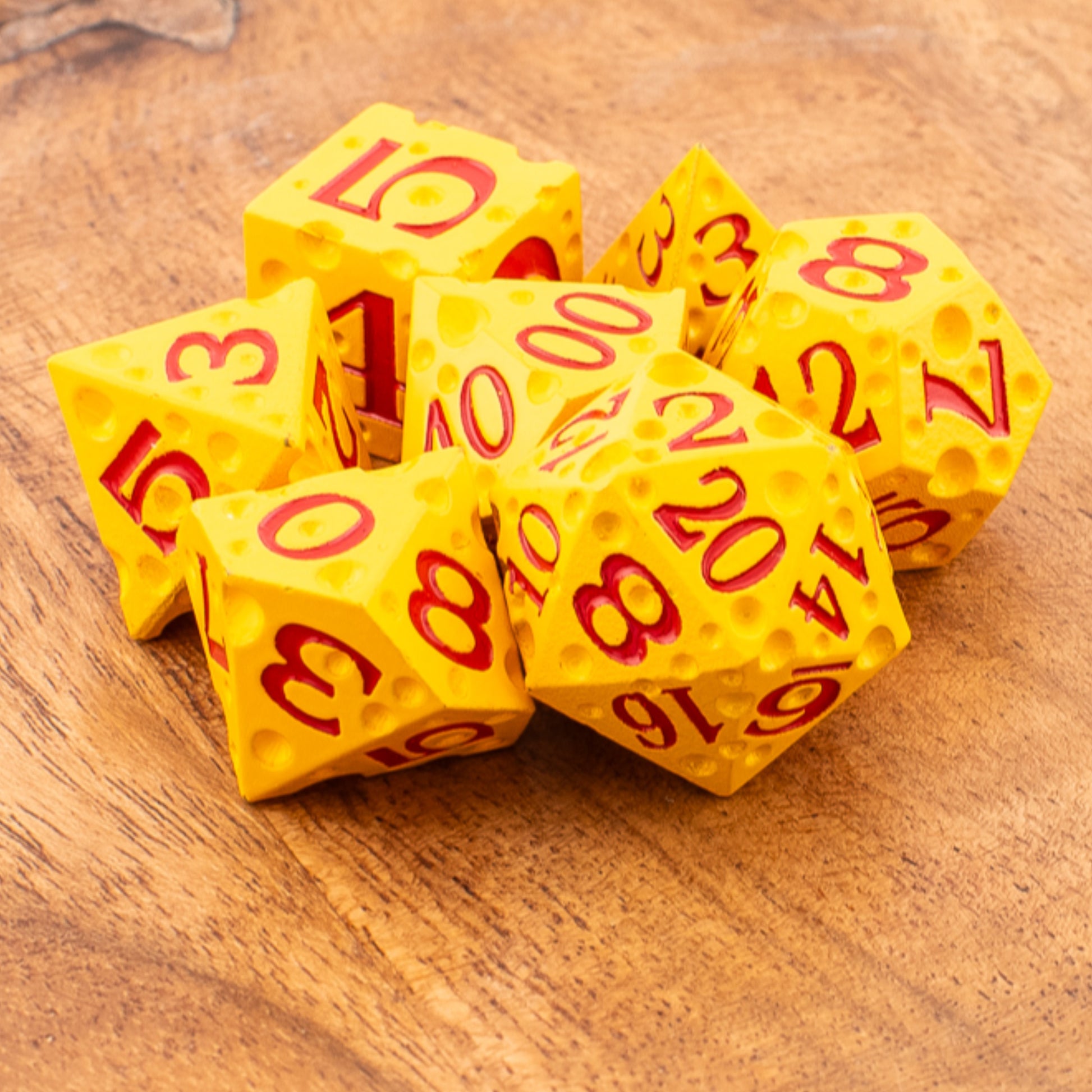 Tabletop Dominion Cheddar Champion heavy metal dice shown loose on a wooden surface, designed with a cheese‑like texture in yellow with red numbering.