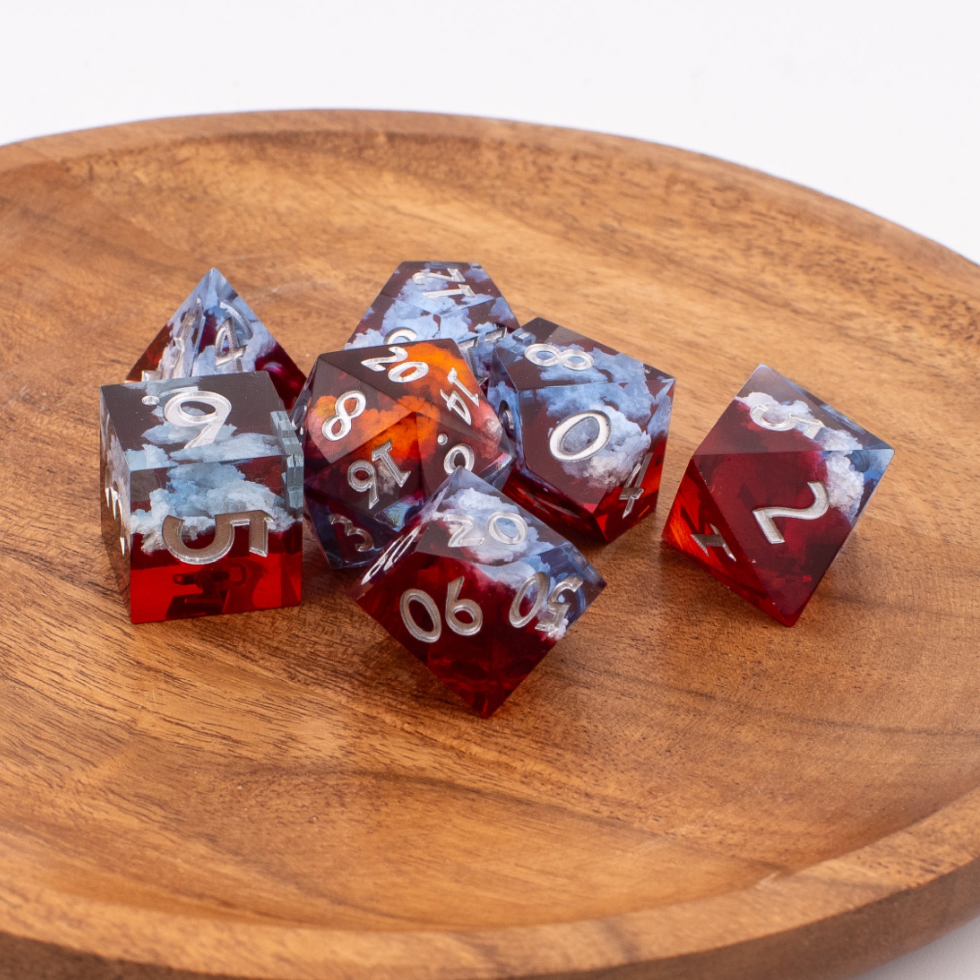 Ember Cloud resin dice scattered on a wooden surface, contrasting the warm wood grain with the red‑and‑blue cloud inclusions inside each sharp‑edge die.